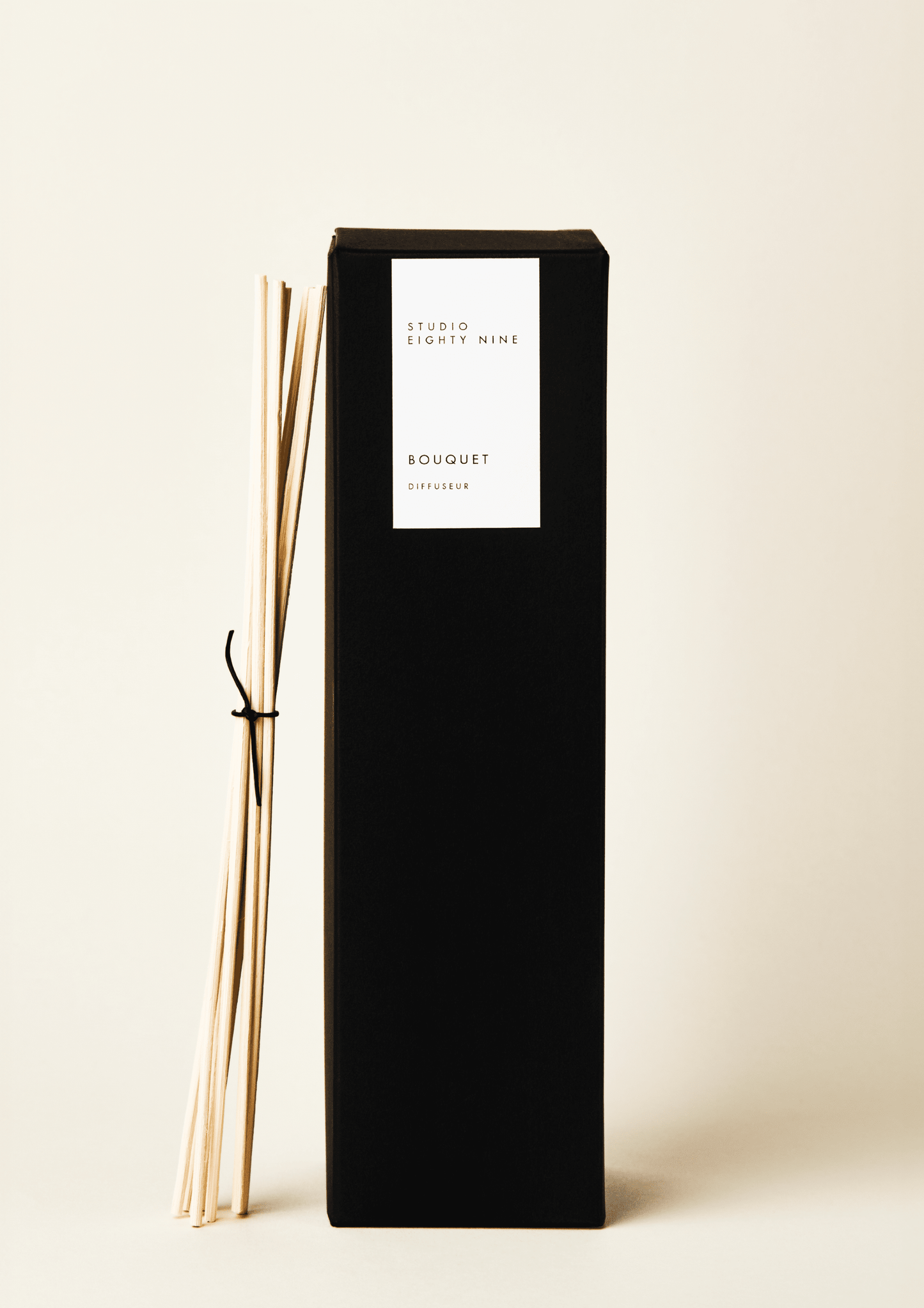 Tall black rectangular diffuser box with a white minimalist rectangular vertical label on the front, featuring the brand 'STUDIO EIGHTY NINE', scent 'BOUQUET', and product 'DIFFUSEUR' in clean, uppercase typography. Bundled, beige-toned reed diffuser sticks, tied with a thin black leather cord, lean against the side of the diffuser box.
