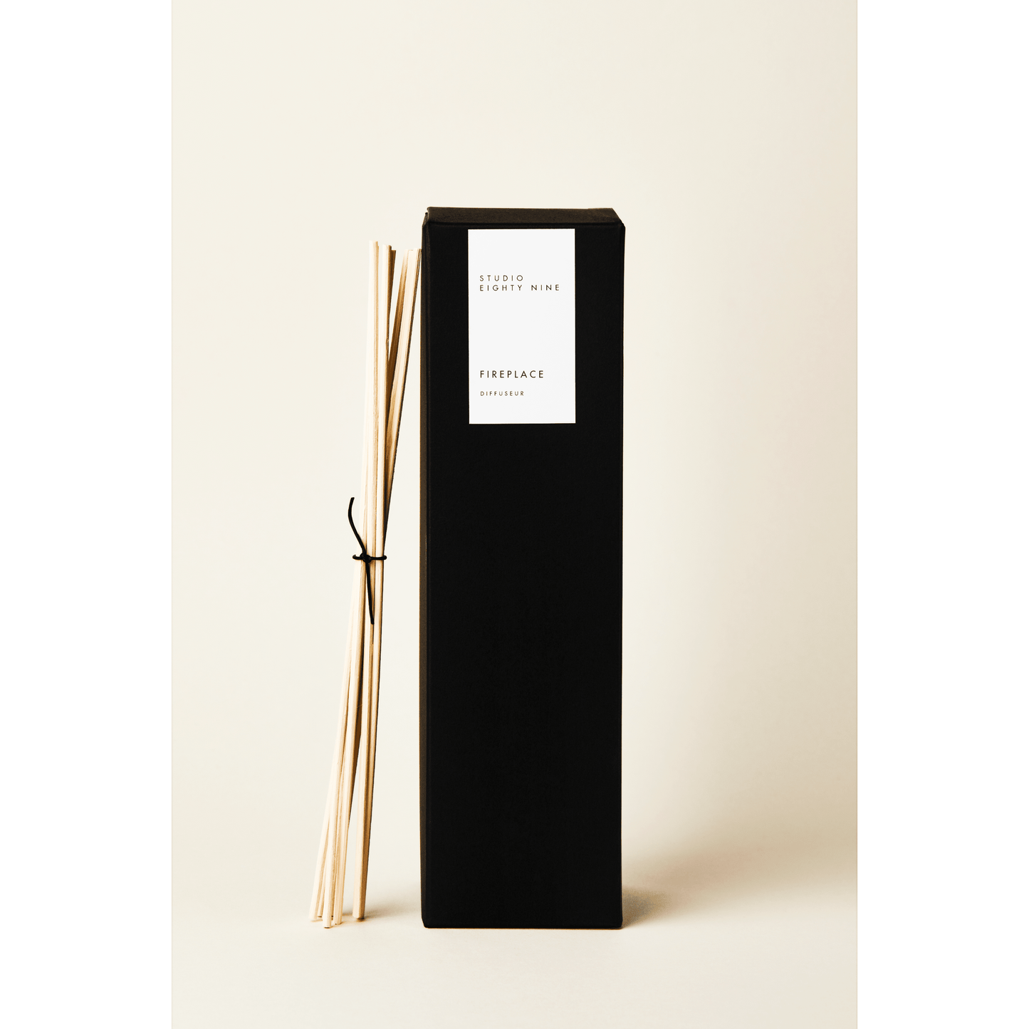 Tall black rectangular diffuser box with a white minimalist rectangular vertical label on the front, featuring the brand 'STUDIO EIGHTY NINE', scent 'FIREPLACE', and product 'DIFFUSEUR' in clean, uppercase typography. Bundled, beige-toned reed diffuser sticks, tied with a thin black leather cord, lean against the side of the diffuser box.