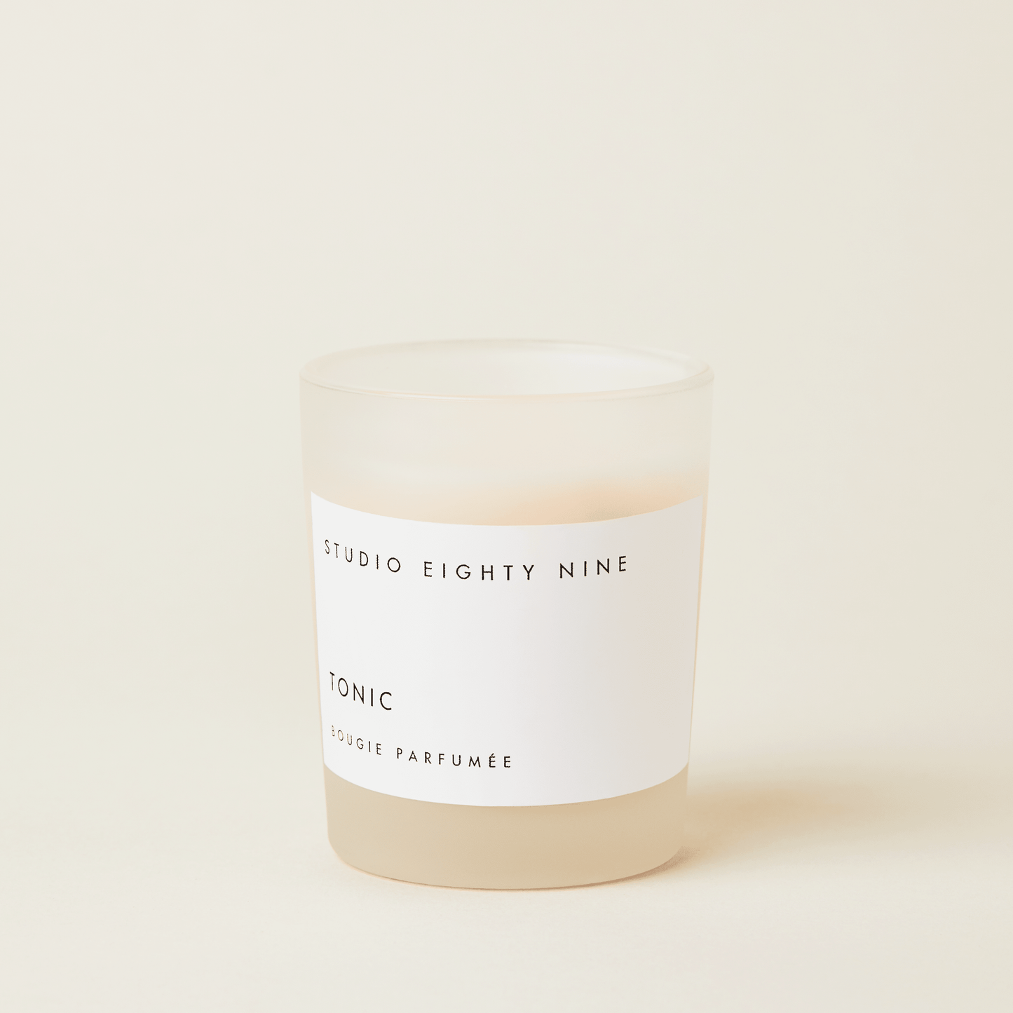 Petite-sized scented candle in frosted glass with beige-toned wax, featuring a white, minimalist rectangular horizontal label showcasing the brand 'STUDIO EIGHTY NINE', scent 'TONIC', and product 'BOUGIE PARFUMÉE' in clean uppercase typography.