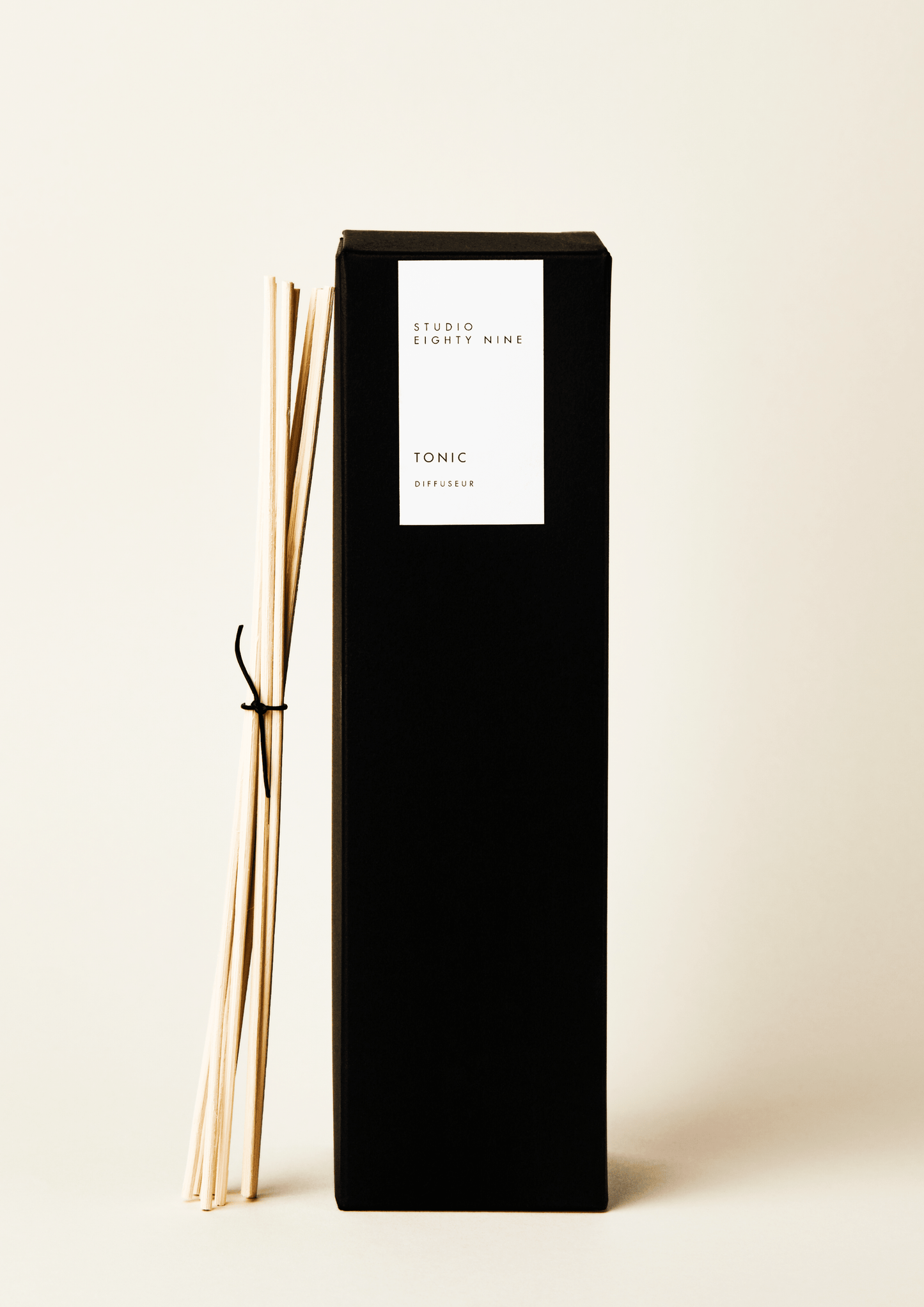 Tall black rectangular diffuser box with a white minimalist rectangular vertical label on the front, featuring the brand 'STUDIO EIGHTY NINE', scent 'TONIC', and product 'DIFFUSEUR' in clean, uppercase typography. Bundled, beige-toned reed diffuser sticks, tied with a thin black leather cord, lean against the side of the diffuser box.