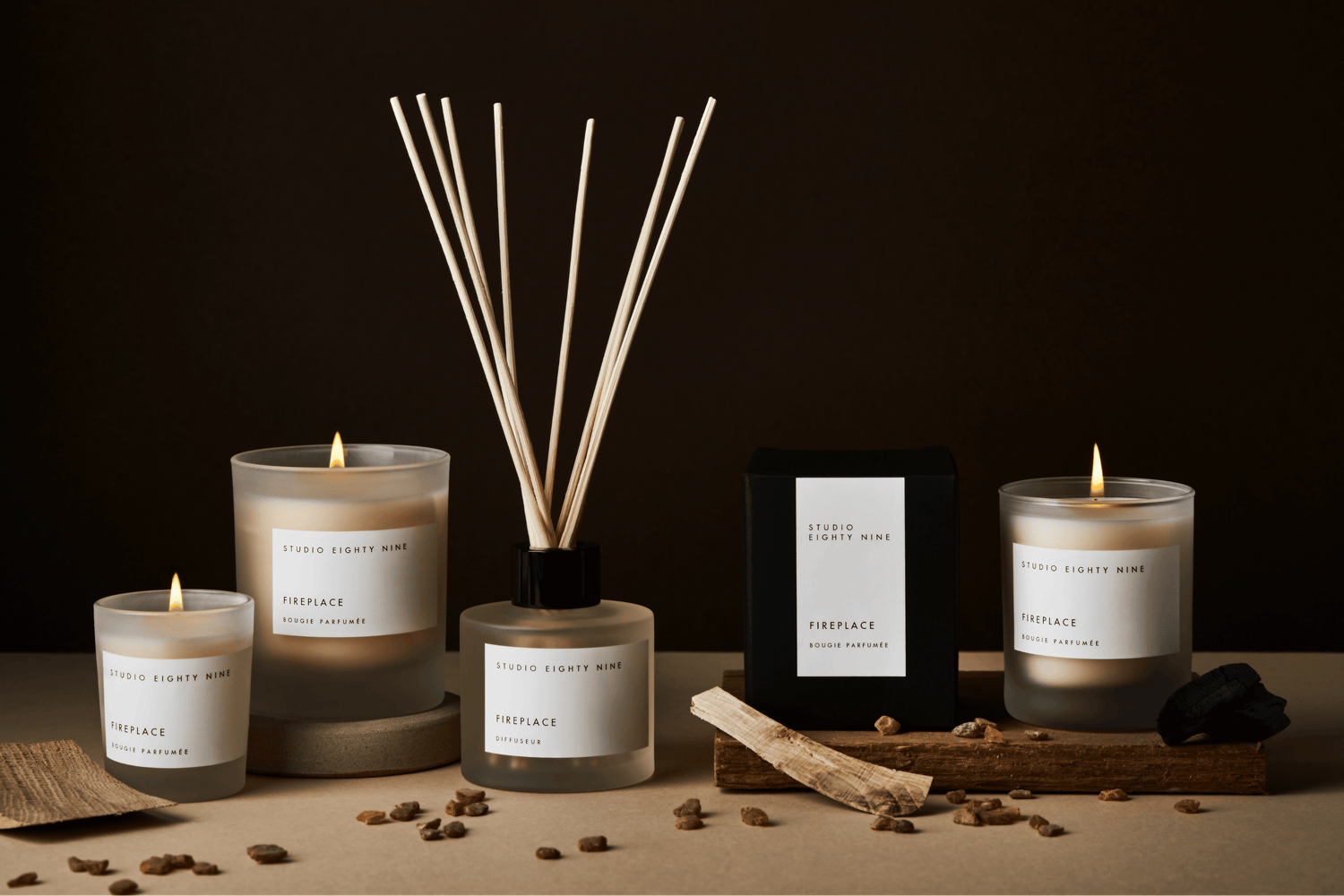 Studio Eighty Nine Fireplace fragrance collection displayed on a dark brown background. The collection includes petite, classic and grande-sized scented candles, and a squat circular reed diffuser, in frosted glass, featuring white minimalist labels with scent ‘FIREPLACE’ in clean, uppercase typography. Pieces of amber, wood and charcoal are surround the products. 