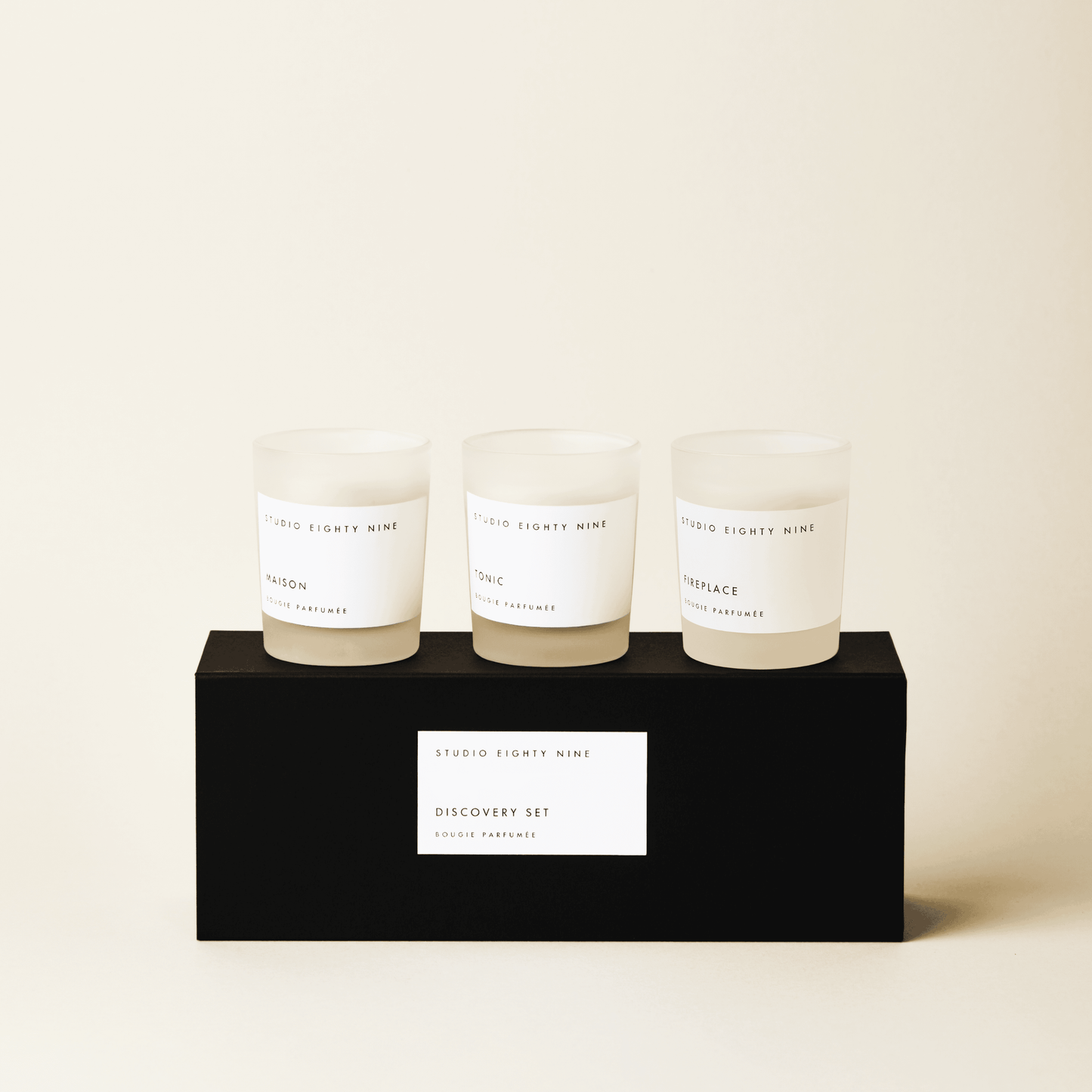 Black rectangular discovery set box featuring a white, minimalist rectangular horizontal label featuring the brand 'STUDIO EIGHTY NINE', description 'DISCOVERY SET', and product 'BOUGIE PARFUMÉE' in clean uppercase typography. Atop the box are three petite-sized scented candles in frosted glass with beige-toned wax. The three featured scents are 'MAISON', 'TONIC', and ‘FIREPLACE’.
