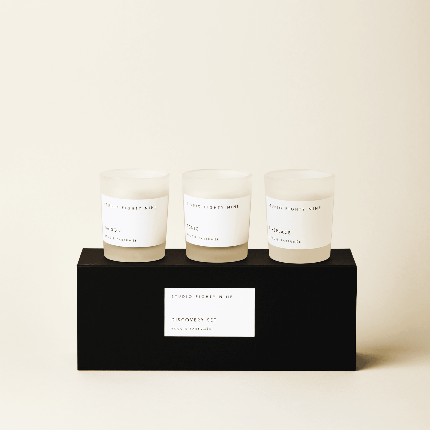 Black rectangular discovery set box featuring a white, minimalist rectangular horizontal label featuring the brand 'STUDIO EIGHTY NINE', description 'DISCOVERY SET', and product 'BOUGIE PARFUMÉE' in clean uppercase typography. Atop the box are three petite-sized scented candles in frosted glass with beige-toned wax. The three featured scents are 'MAISON', 'TONIC', and ‘FIREPLACE’.