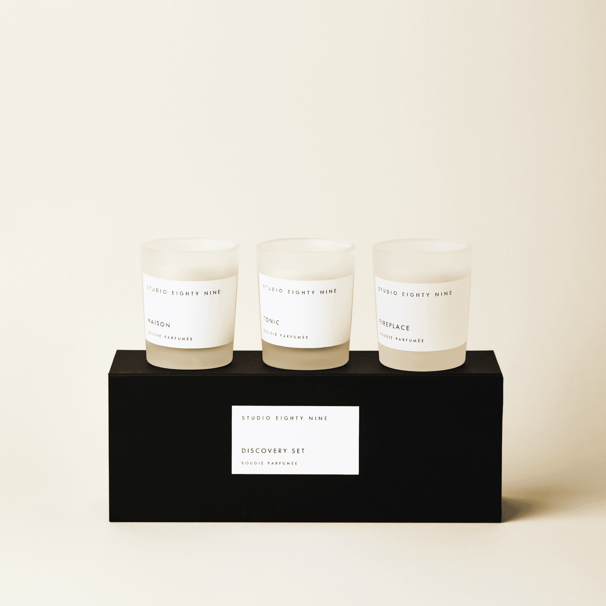 Black rectangular discovery set box featuring a white, minimalist rectangular horizontal label featuring the brand 'STUDIO EIGHTY NINE', description 'DISCOVERY SET', and product 'BOUGIE PARFUMÉE' in clean uppercase typography. Atop the box are three petite-sized scented candles in frosted glass with beige-toned wax. The three featured scents are 'MAISON', 'TONIC', and ‘FIREPLACE’.