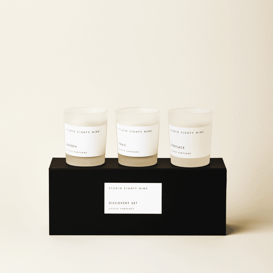 Black rectangular discovery set box featuring a white, minimalist rectangular horizontal label featuring the brand 'STUDIO EIGHTY NINE', description 'DISCOVERY SET', and product 'BOUGIE PARFUMÉE' in clean uppercase typography. Atop the box are three petite-sized scented candles in frosted glass with beige-toned wax. The three featured scents are 'MAISON', 'TONIC', and ‘FIREPLACE’.
